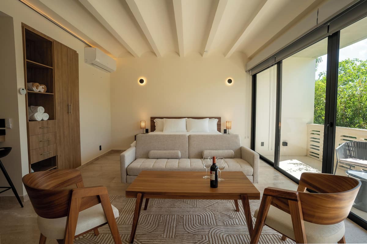 Quarto moderno com sofá-cama, uma mesa de madeira com uma garrafa e uma janela grande com vista para o verde.