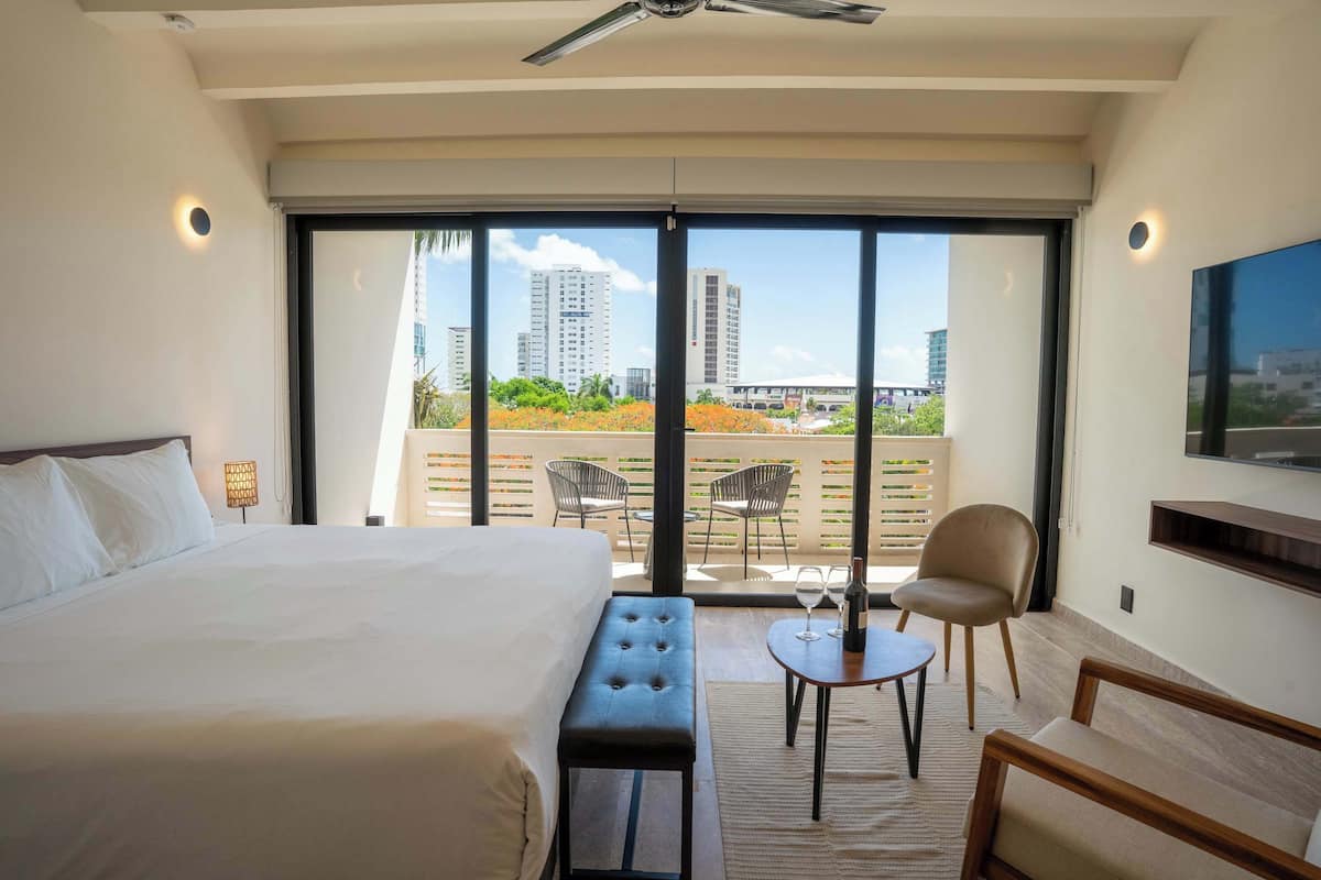 Quarto de hotel com uma cama grande, um banco, uma mesa pequena com uma taça de vinho e uma cadeira. O quarto tem uma varanda com vista para edifícios e áreas verdes.