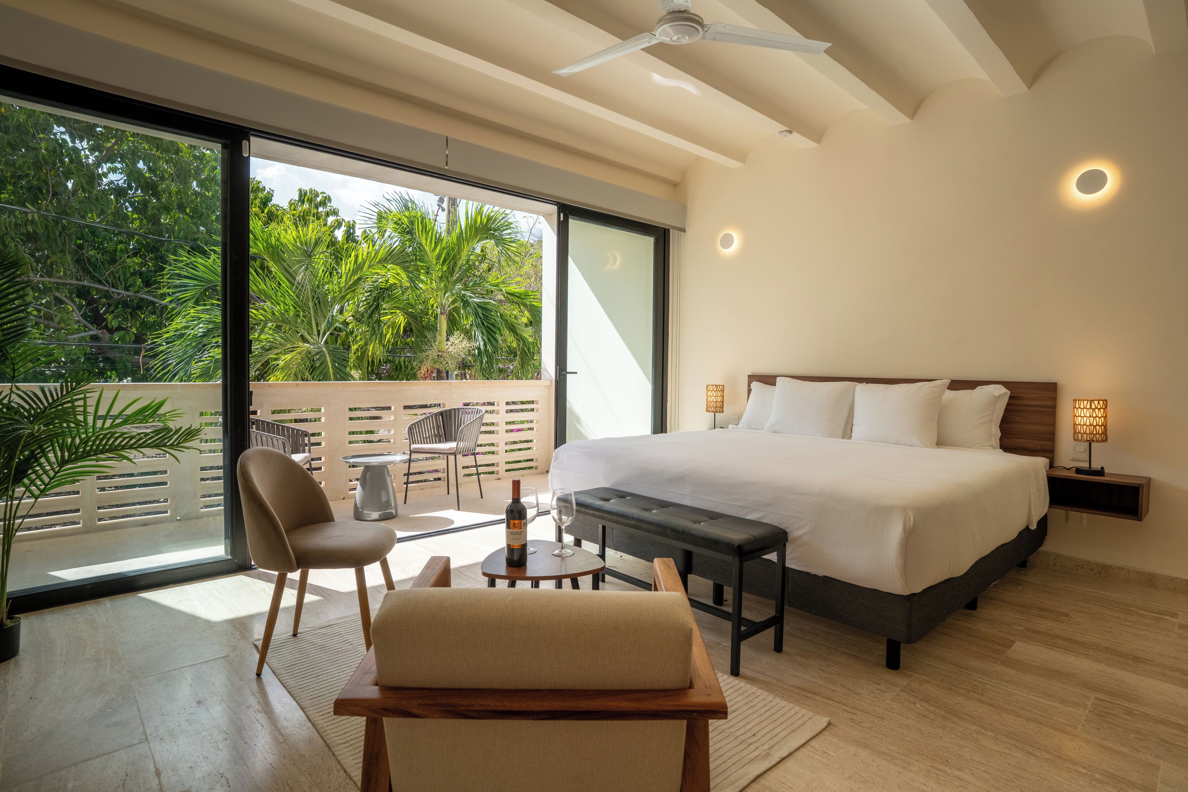 A modern bedroom with a large bed, a seating area, and a balcony with a view of lush greenery.