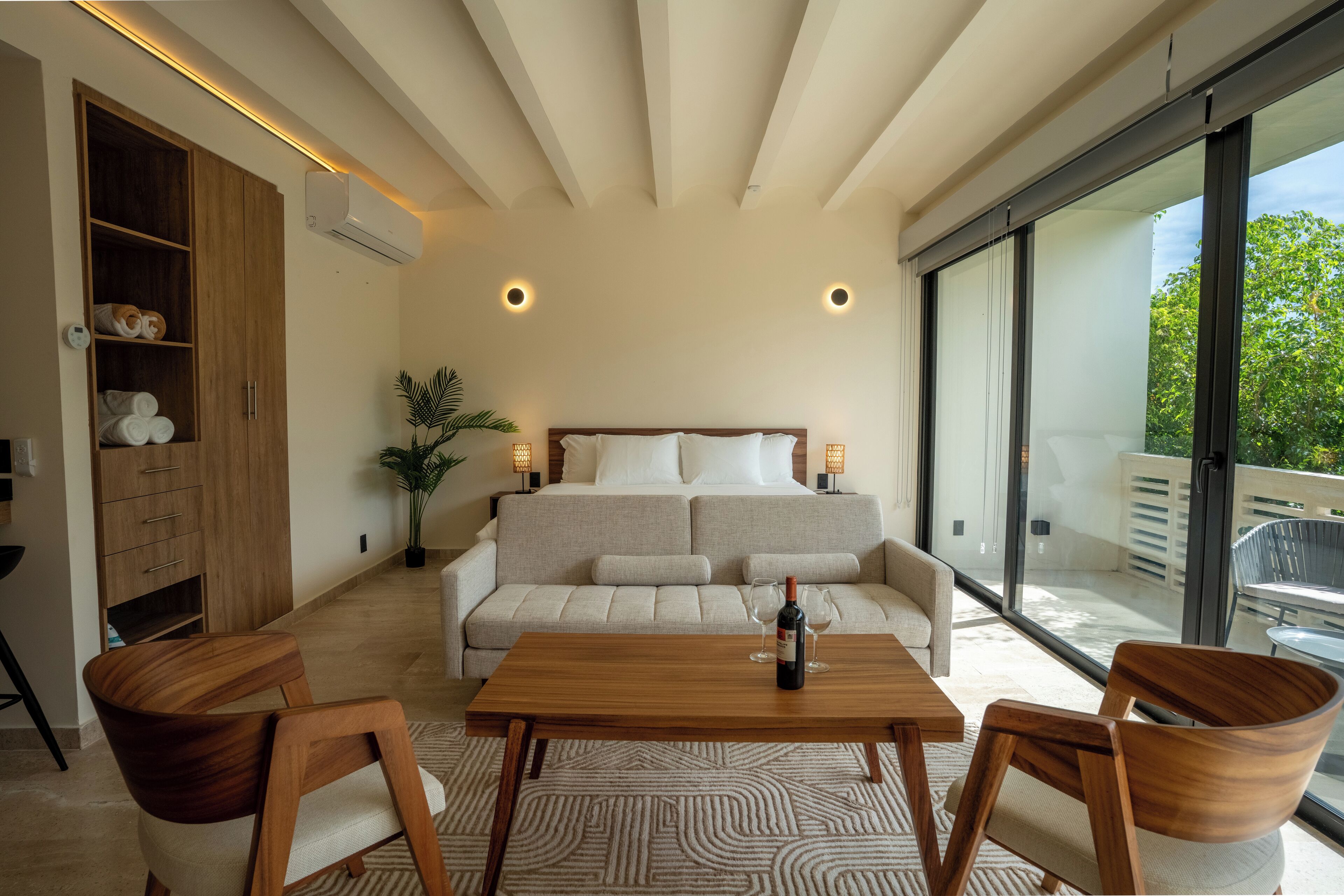 A modern bedroom with a large bed, a wooden wardrobe, a dining table, and a view of the outdoors.