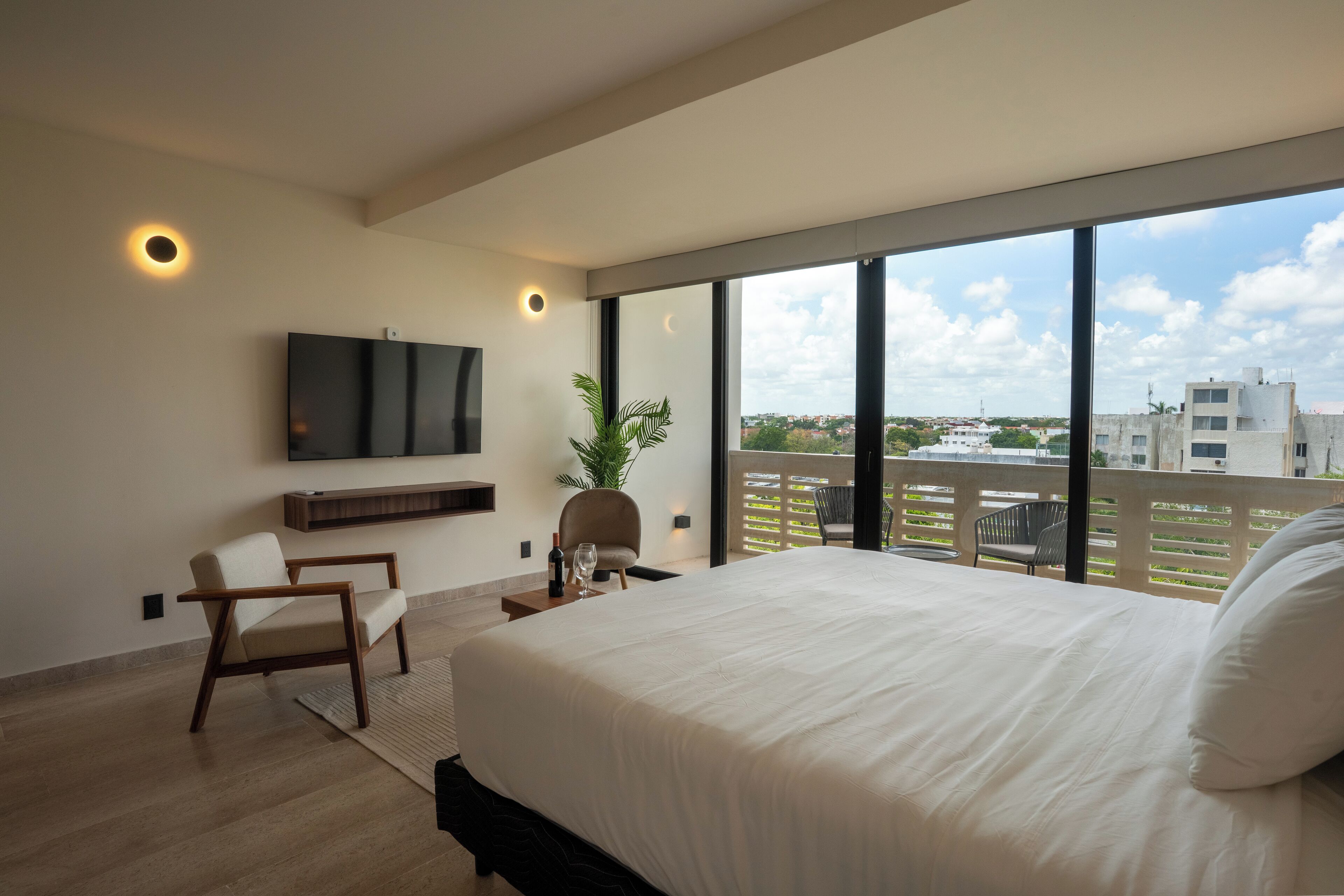 A modern hotel room with a large bed, a TV mounted on the wall, a potted plant, and a balcony with a view of the city.