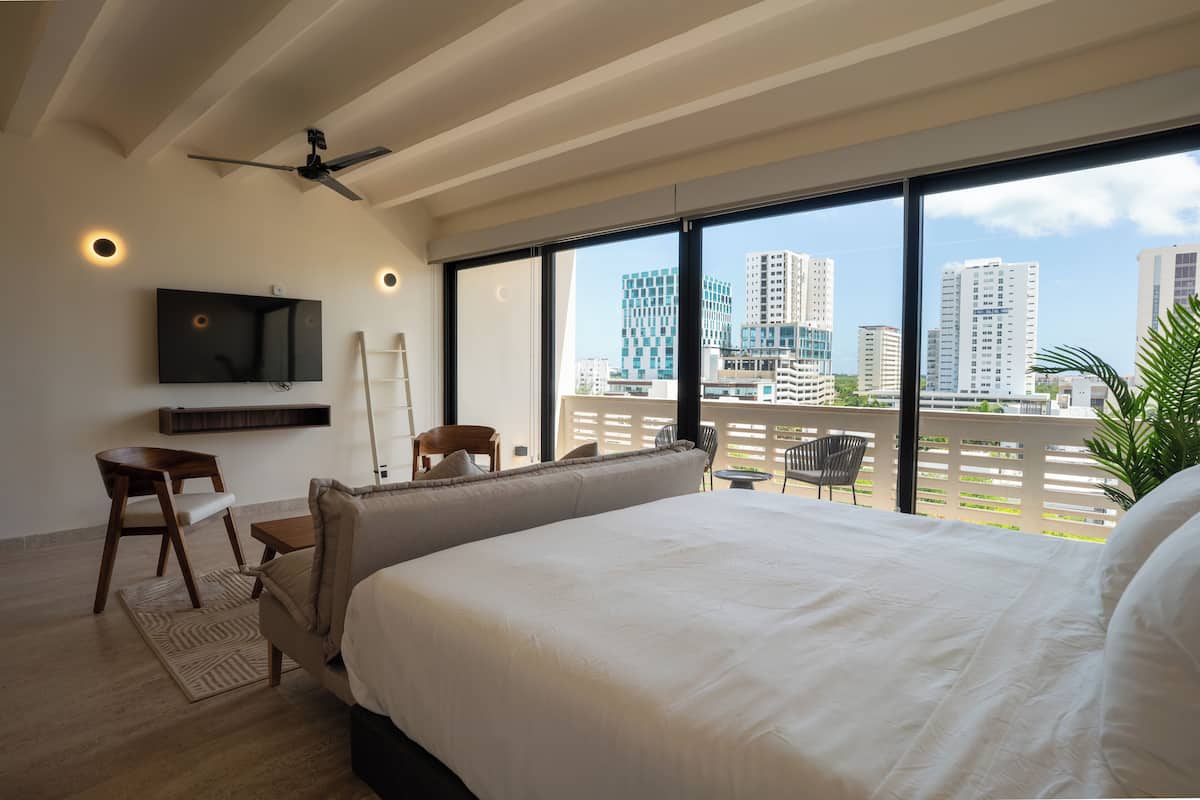 Quarto de hotel moderno com uma cama grande, televisão na parede, ventilador de teto e varanda com vista para a cidade.