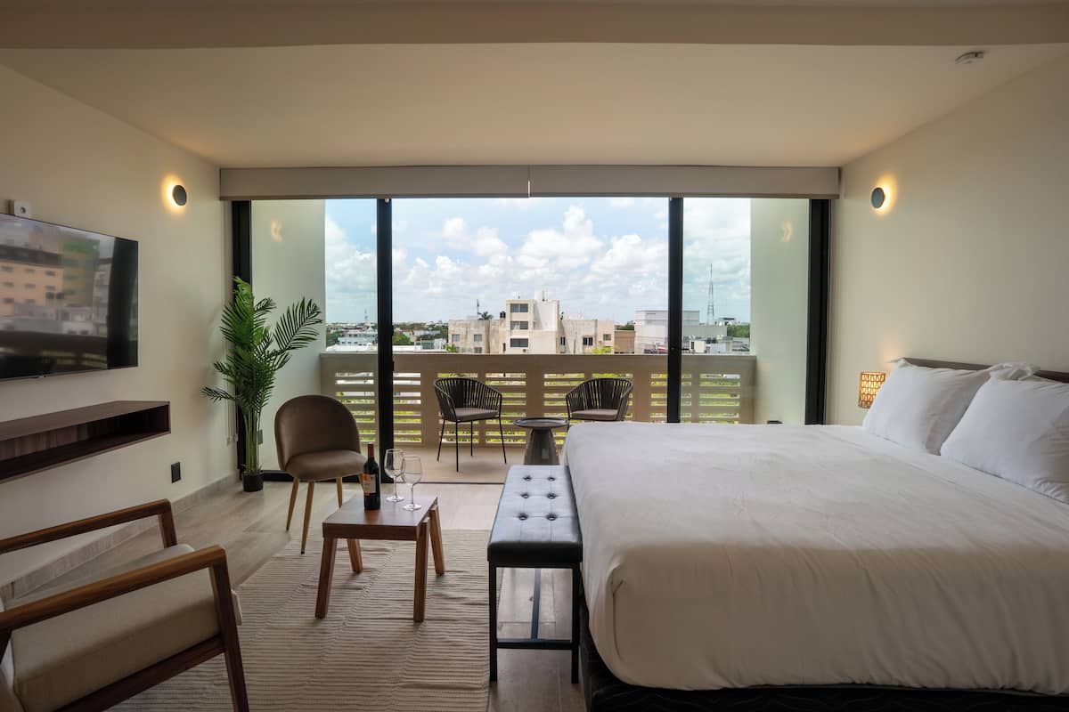 Quarto de hotel moderno com uma cama grande, televisão, varanda com assentos e vista para a cidade.