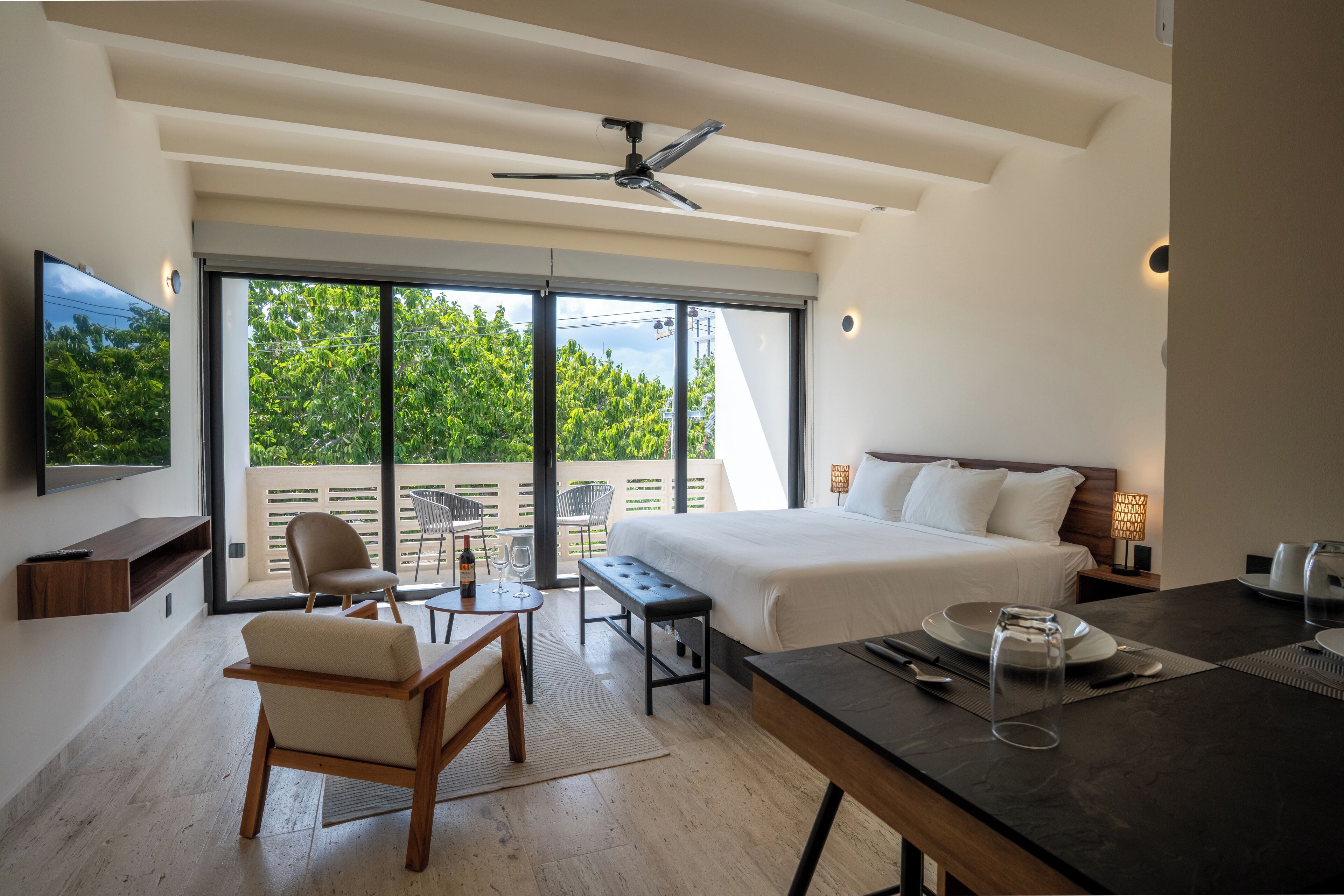 A modern hotel room with a large bed, a dining table, a sofa, a TV, and a balcony with a view of trees.