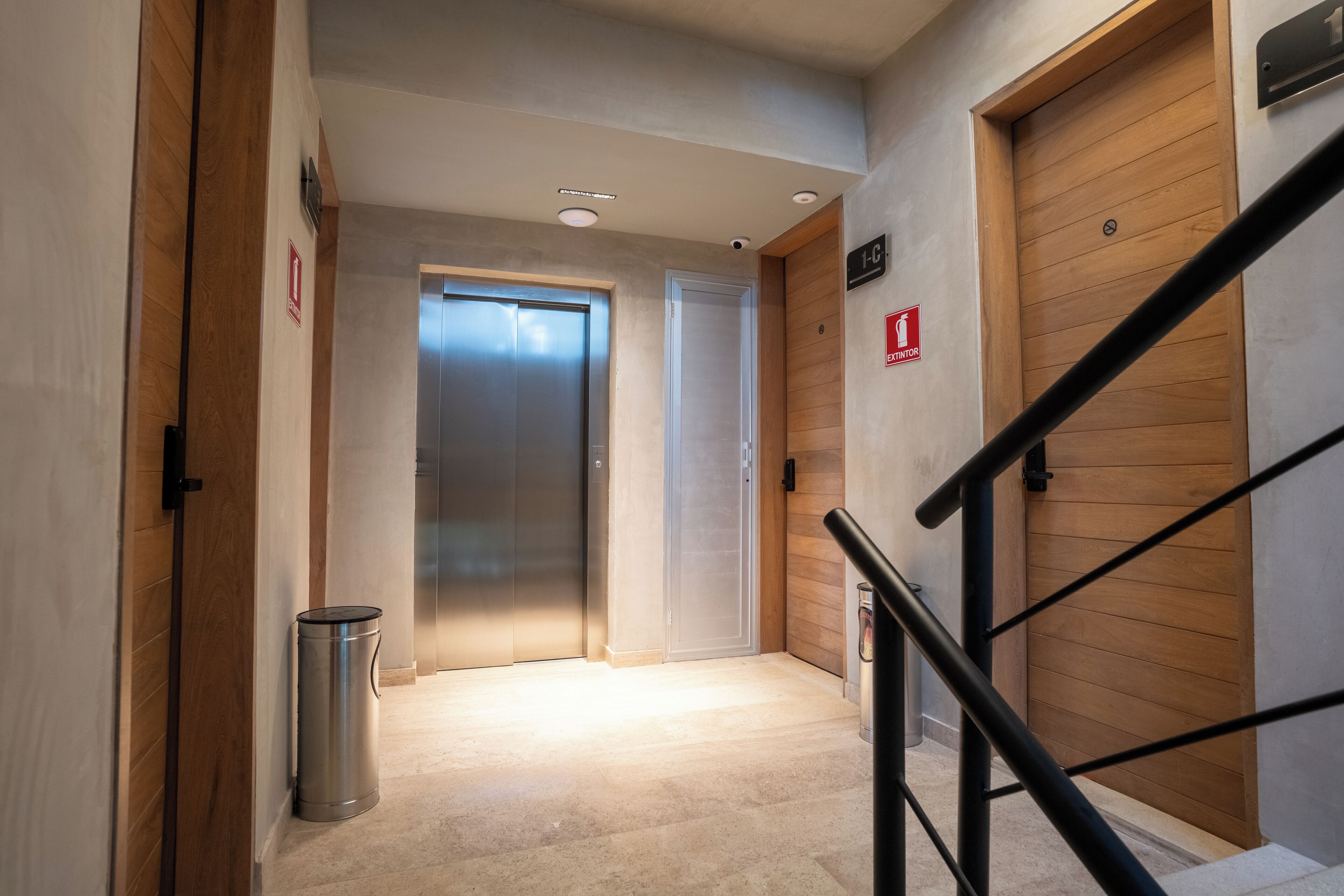 A hallway with a trash bin, two doors, a staircase, and a fire exit sign.