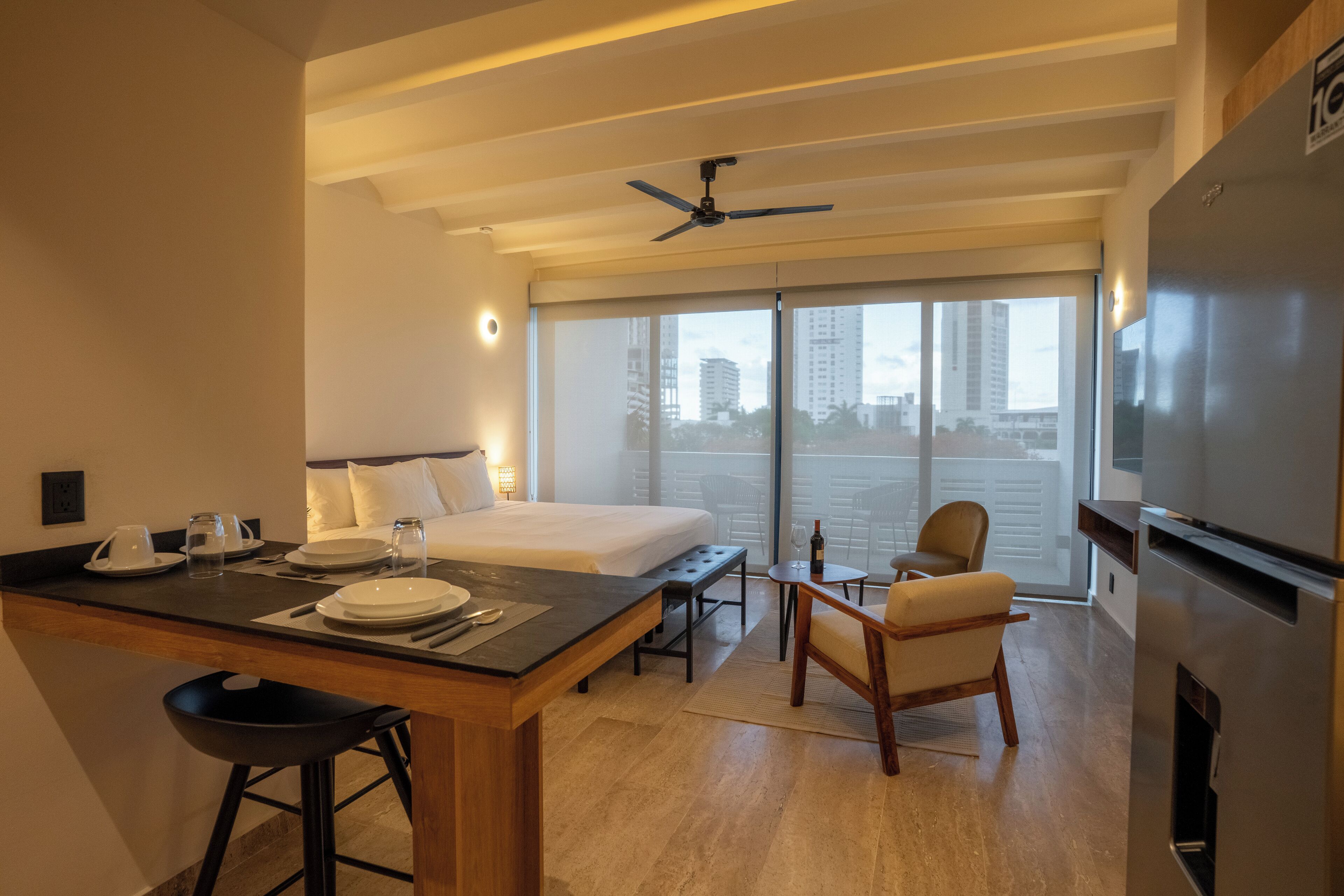 Ein modernes Hotelzimmer mit einem großen Bett, einem Essbereich mit Tisch und Stühlen, einer Küche inklusive Kühlschrank und Blick auf die Stadt durch große Fenster.