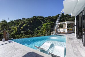 A swimming pool with a waterfall feature, surrounded by lush greenery and a modern outdoor seating area.