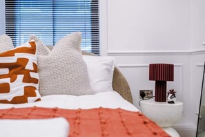 Quarto com cama, almofadas, abajur, uma planta pequena e janela com persianas.