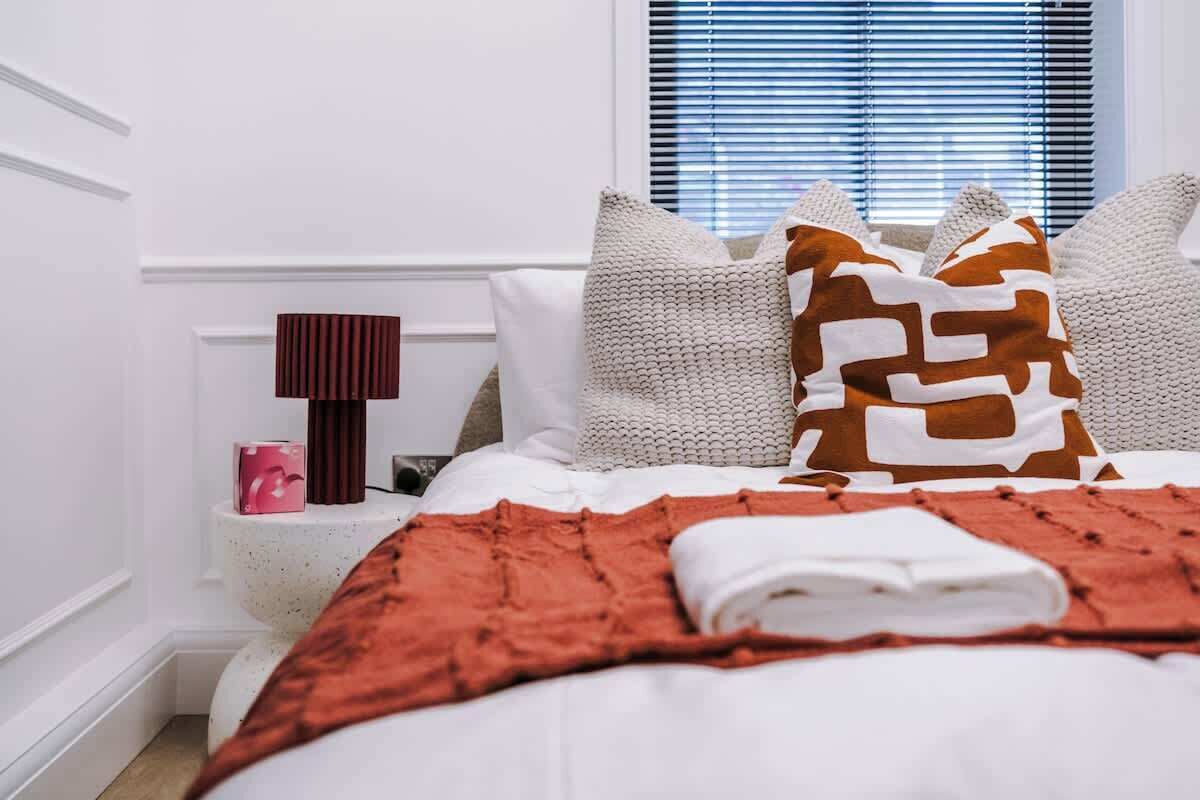 Un lit bien fait, avec une couverture rouge, des oreillers blancs et un coussin à motifs bruns.