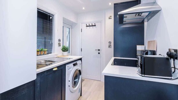 A compact kitchen with a washing machine, a toaster, and a small window with blinds. - Stylish 1-Bed Flat | York Centre | Sleeps 4 (York)