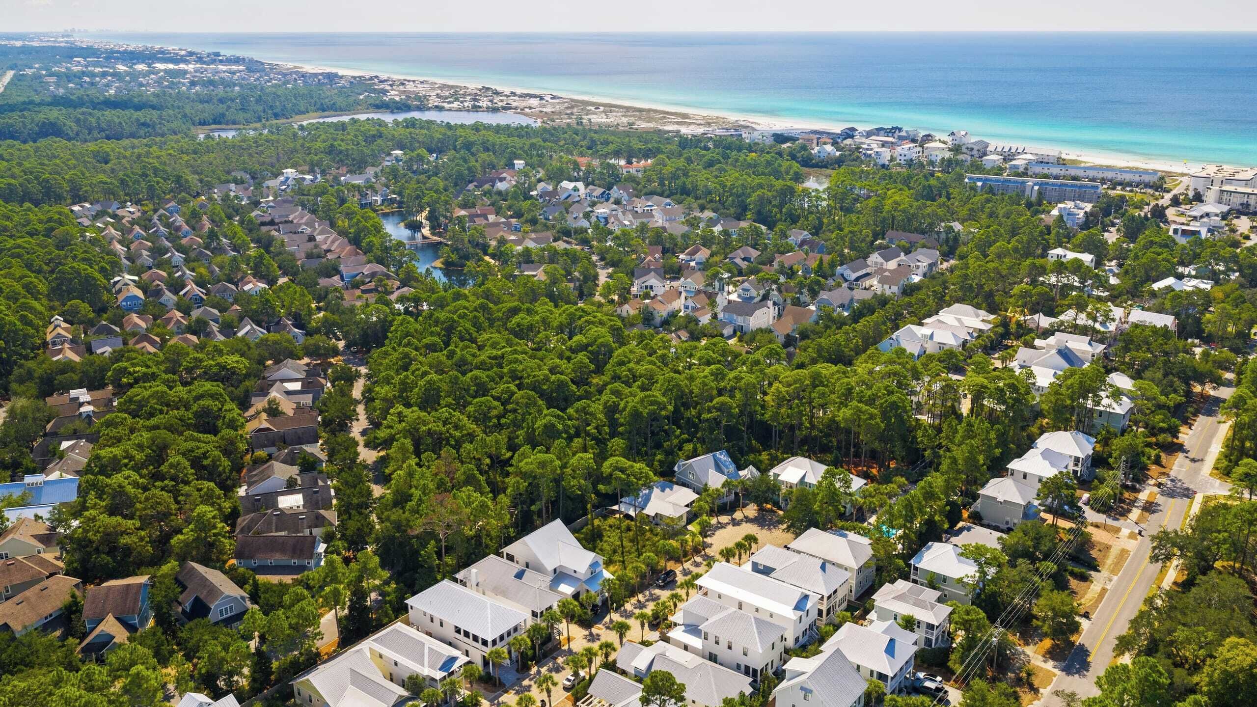 Vue aérienne d’un quartier résidentiel avec des maisons, une végétation luxuriante et une plage en arrière-plan.