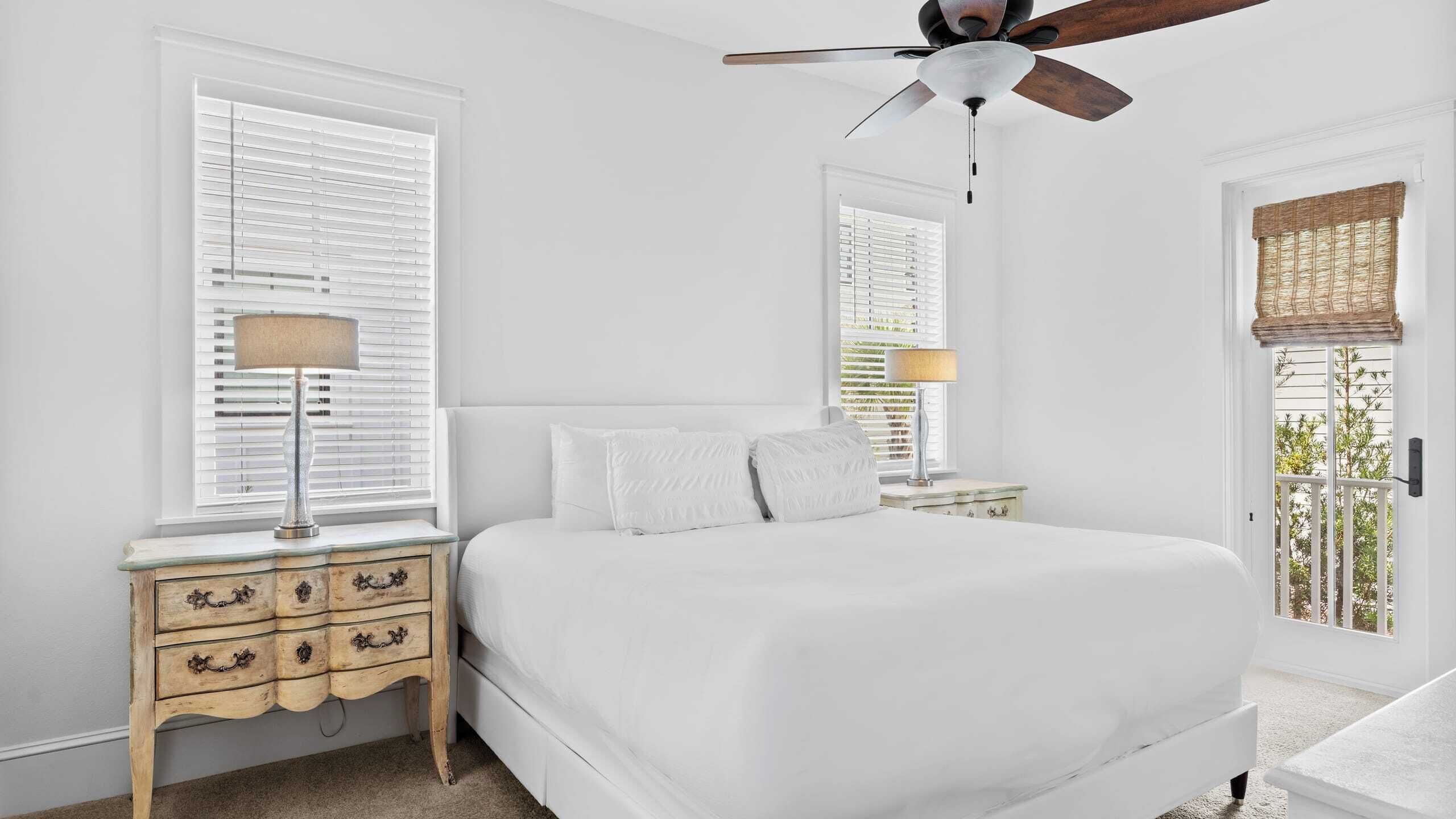 A bedroom with a large bed, two bedside tables with lamps, a ceiling fan, and a window with blinds.