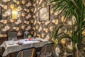 A dining area with a table set for two, chairs, a potted plant, and a floral wallpaper.