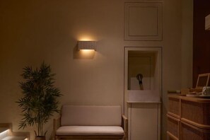 Huoneessa on sohva, seinään kiinnitetty valaisin, kehystetty taulu ja ruukussa oleva kasvi.