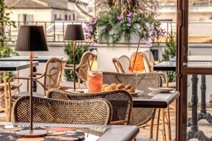 Un espace extérieur aménagé avec des chaises et des tables en osier, une boisson posée sur la table et une vue sur les bâtiments en arrière-plan.