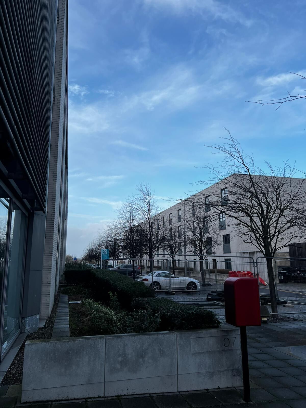 Een straatbeeld met een modern gebouw, een geparkeerde auto en een rode brievenbus.