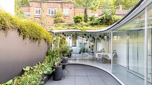 A modern patio with a curved glass wall, a seating area with tables and chairs, and a variety of potted plants. - Listed Georgian Home w/ Contemporary Architecture (London)
