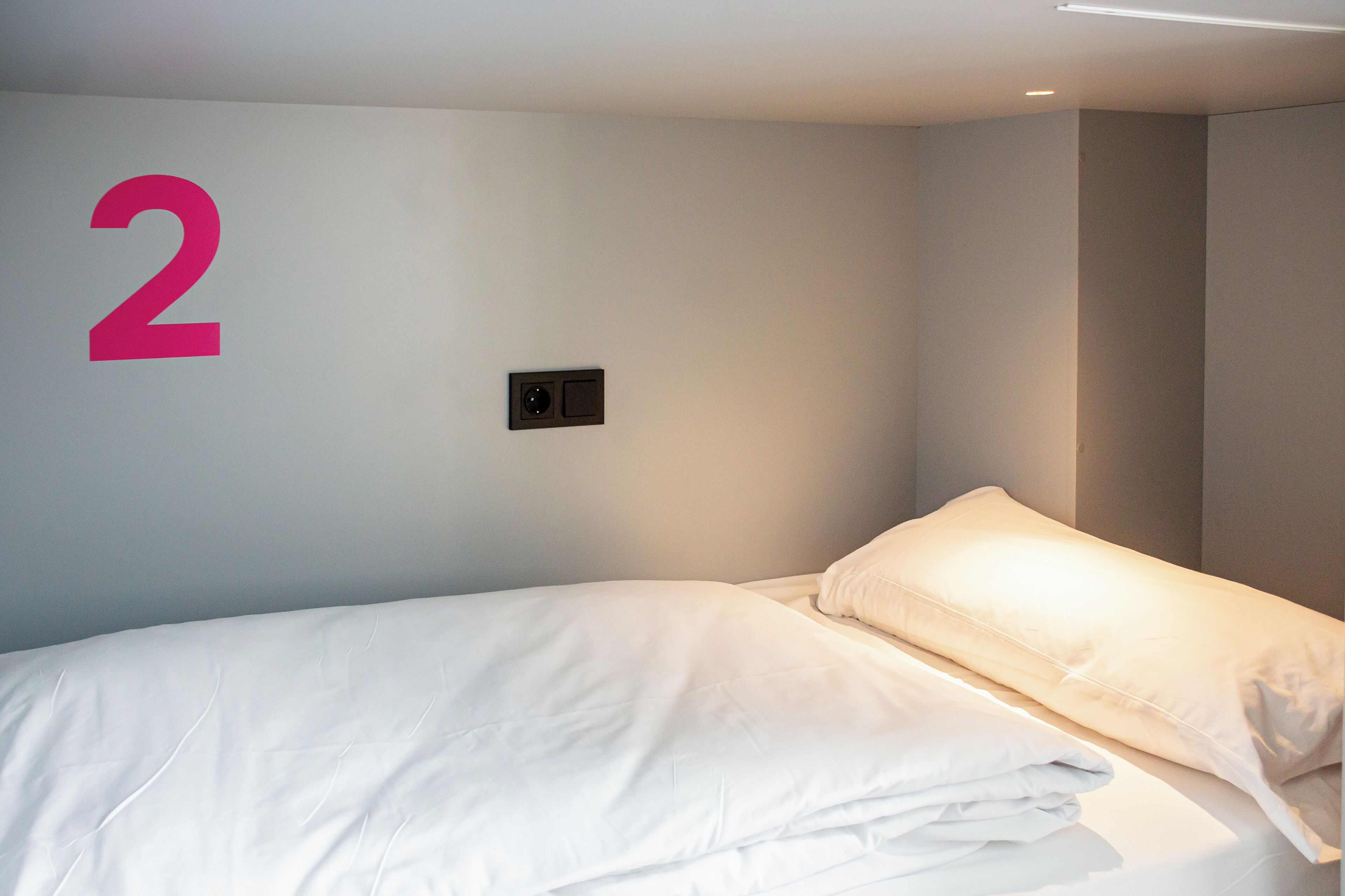 A hotel room with a bed, a wall-mounted light, and a large number '2' on the wall.