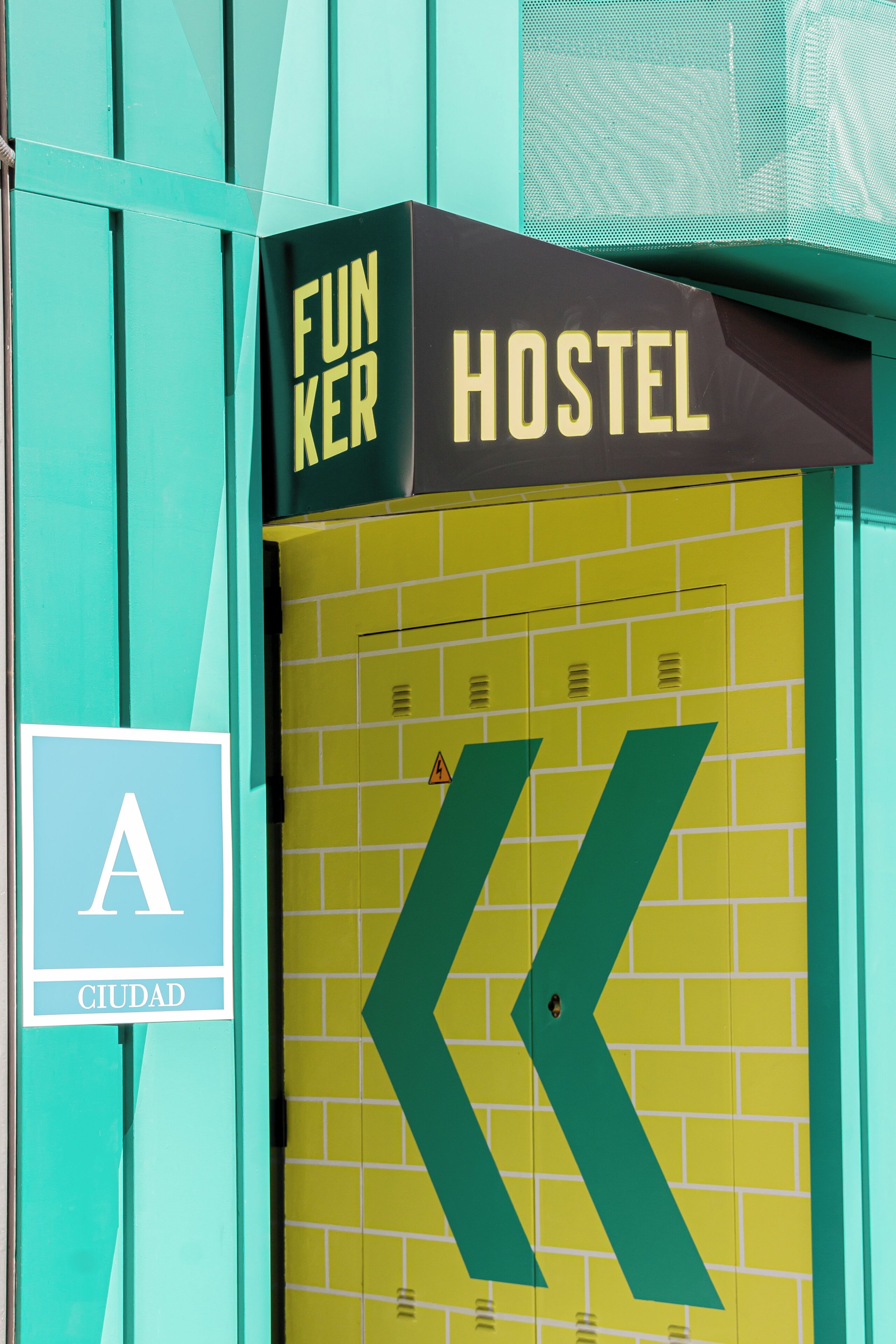 Cartell de l'alberg amb una gran fletxa apuntant a l'esquerra, un rètol 'FUNKER' a la part superior i un logotip 'CIUDAD' sobre un fons color turquesa.