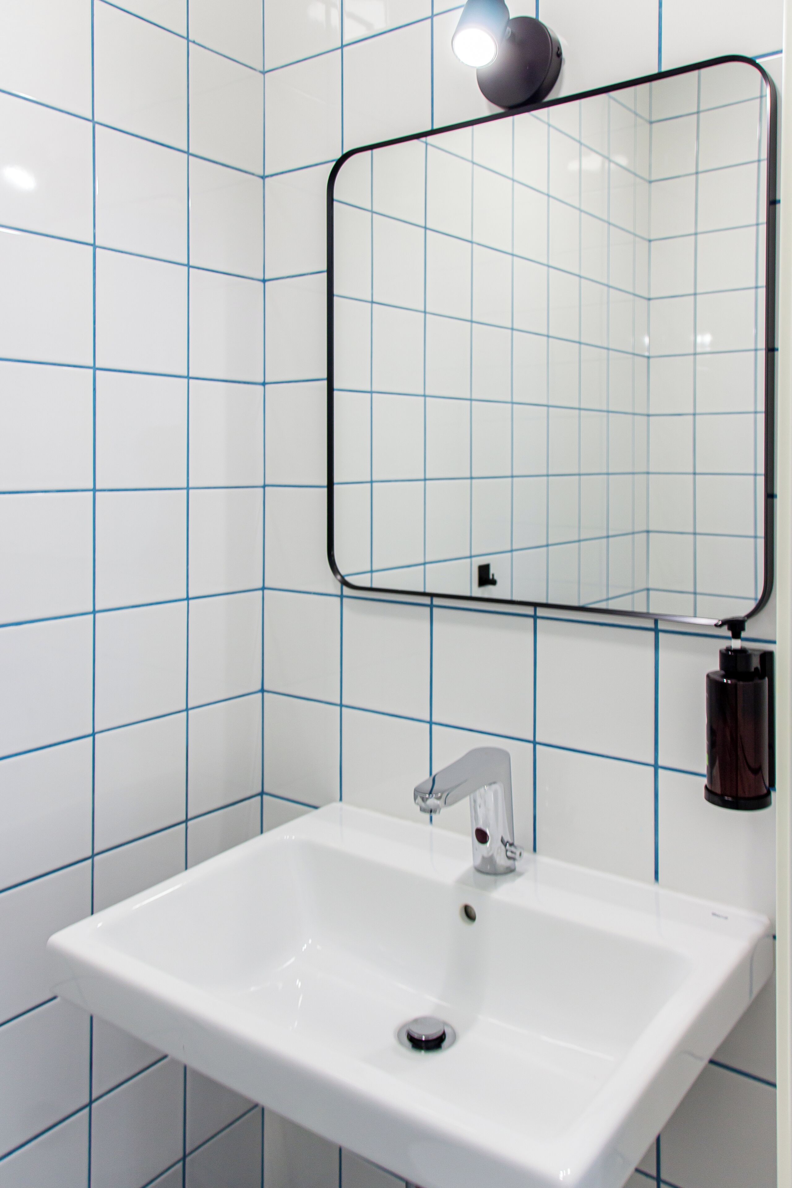 Ein Badezimmer mit weiß gefliesten Wänden, einem rechteckigen Waschbecken mit einem Wasserhahn und einem Spiegel mit schwarzem Rahmen.