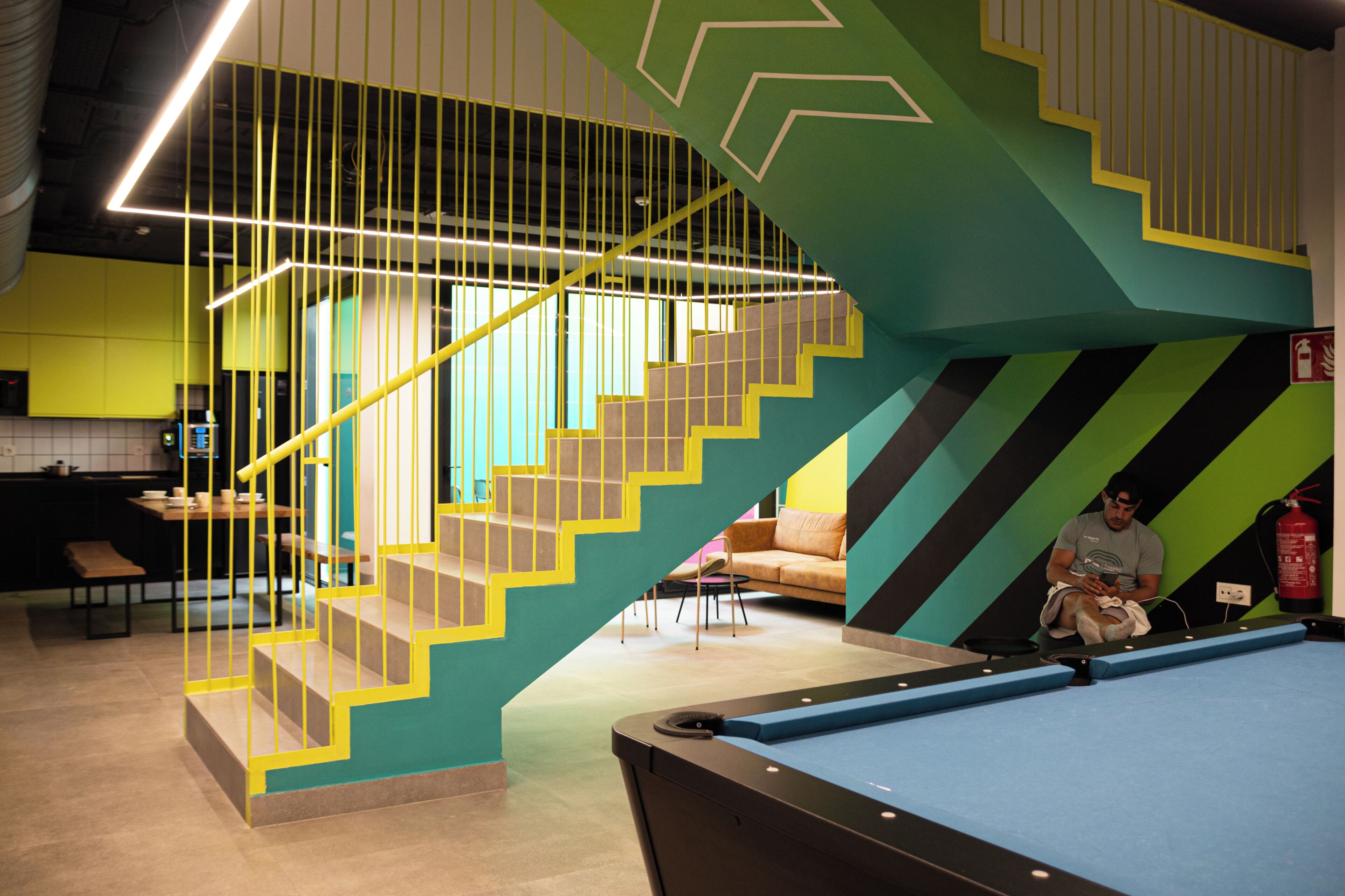Een modern interieur met een pooltafel, een trap met gele leuningen en een brandblusser.