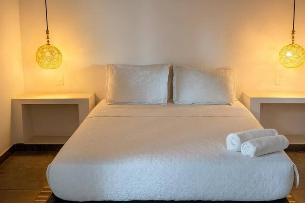 A neatly made bed with pillows and rolled towels, two bedside tables, and hanging lamps.