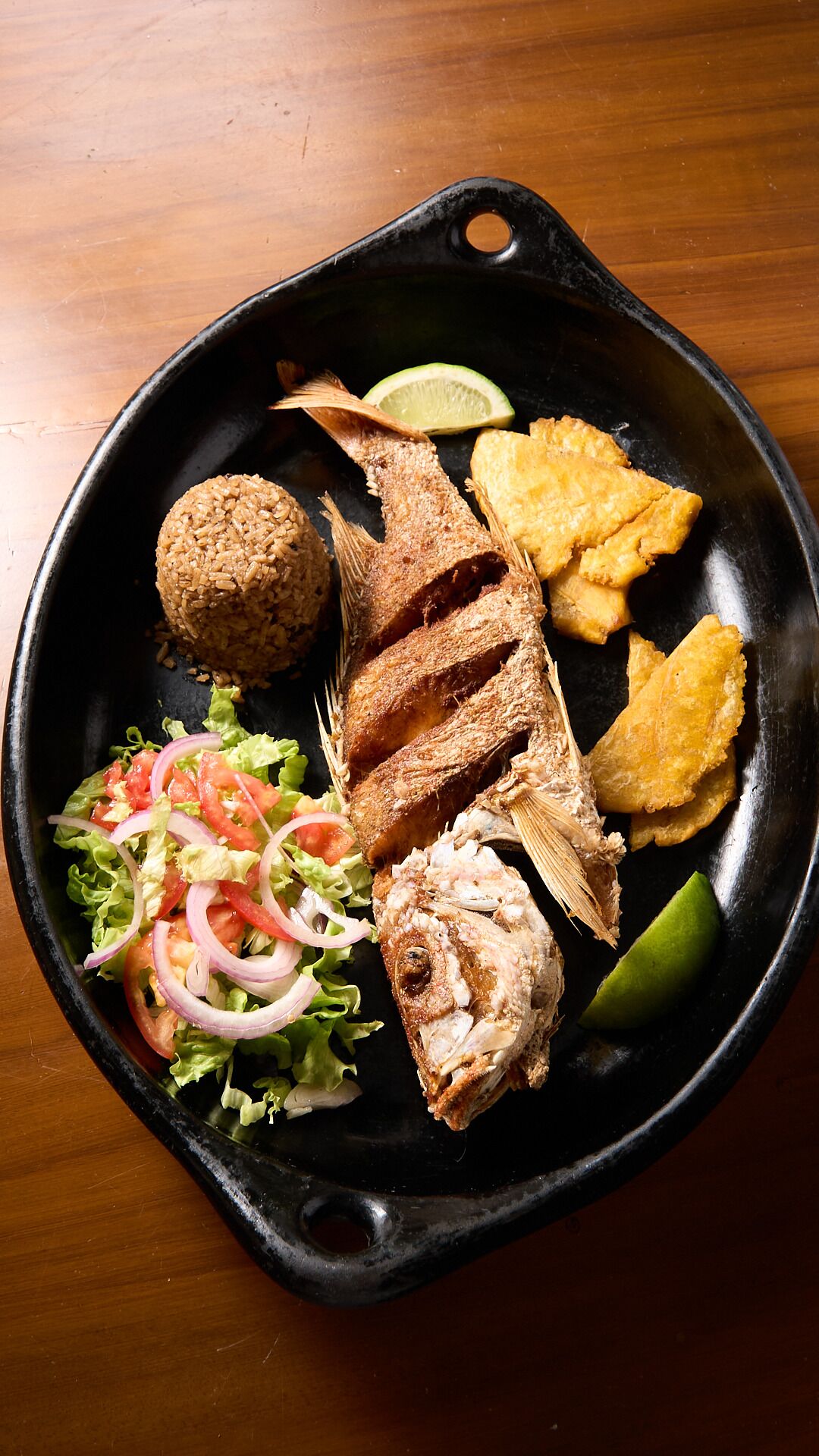 A black skillet with a grilled fish, fried plantains, a rice ball, a salad, and a lime wedge.