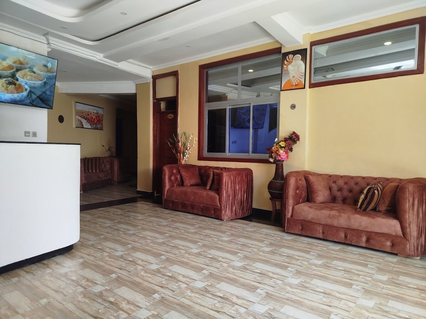 A spacious lobby with a wooden floor, a brown leather sofa, a white reception desk, and a large window.