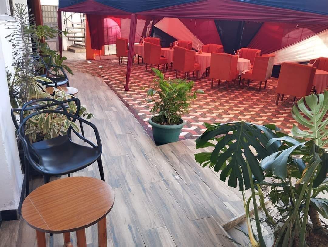 Un espacio al aire libre con mesas y sillas de madera, rodeado de plantas en macetas y un piso con diseño.