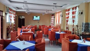 A restaurant with multiple tables and orange chairs, a television on the wall, and red and white curtains.