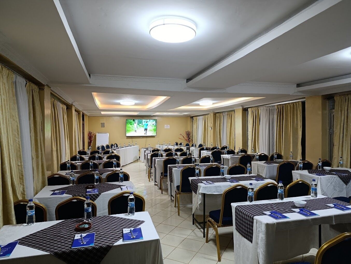 Un salón de conferencias con filas de mesas y sillas, una pantalla grande y una pizarra.