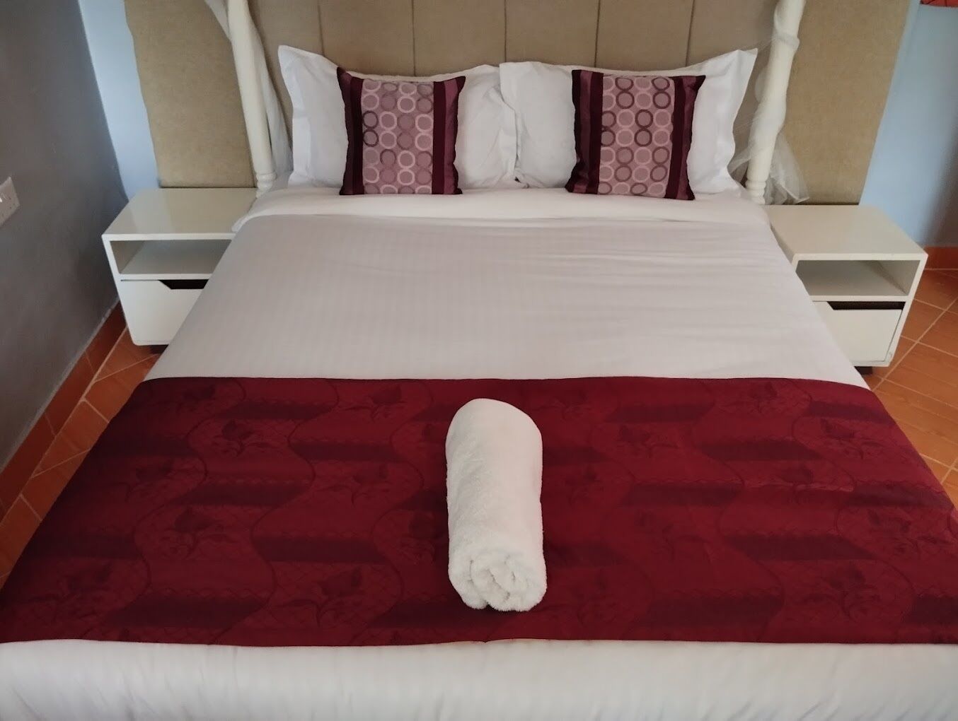 Una cama bien hecha con una colcha roja, una toalla enrollada, almohadas blancas y un cabecero.