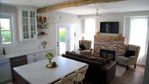 Eine moderne Küche mit weißen Schränken, ein Essbereich mit weißem Tisch und Stühlen, ein Kamin und ein an der Wand befestigter Fernseher.