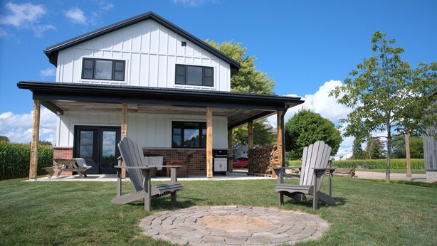 Ein zweistöckiges Haus mit überdachter Terrasse, zwei Adirondack-Stühlen und einer runden Feuerstelle aus Stein.