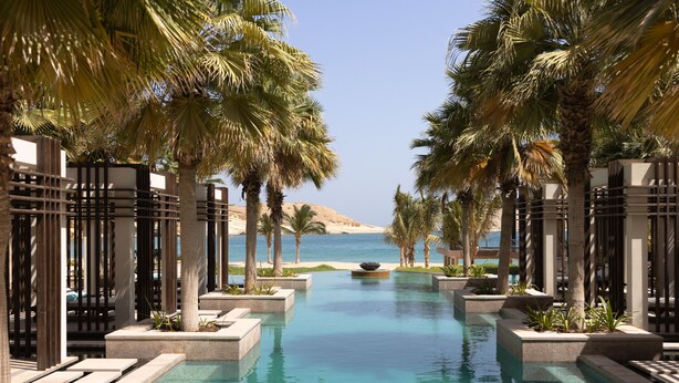A pool with a view of the sea, surrounded by palm trees and a sandy beach. - Jumeirah Muscat Bay (Bandar Jissah)