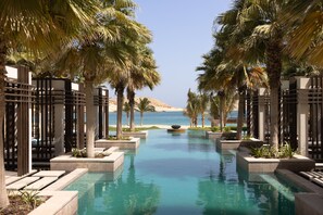 A pool with a view of the sea, surrounded by palm trees and a sandy beach. - Jumeirah Muscat Bay (Bandar Jissah)