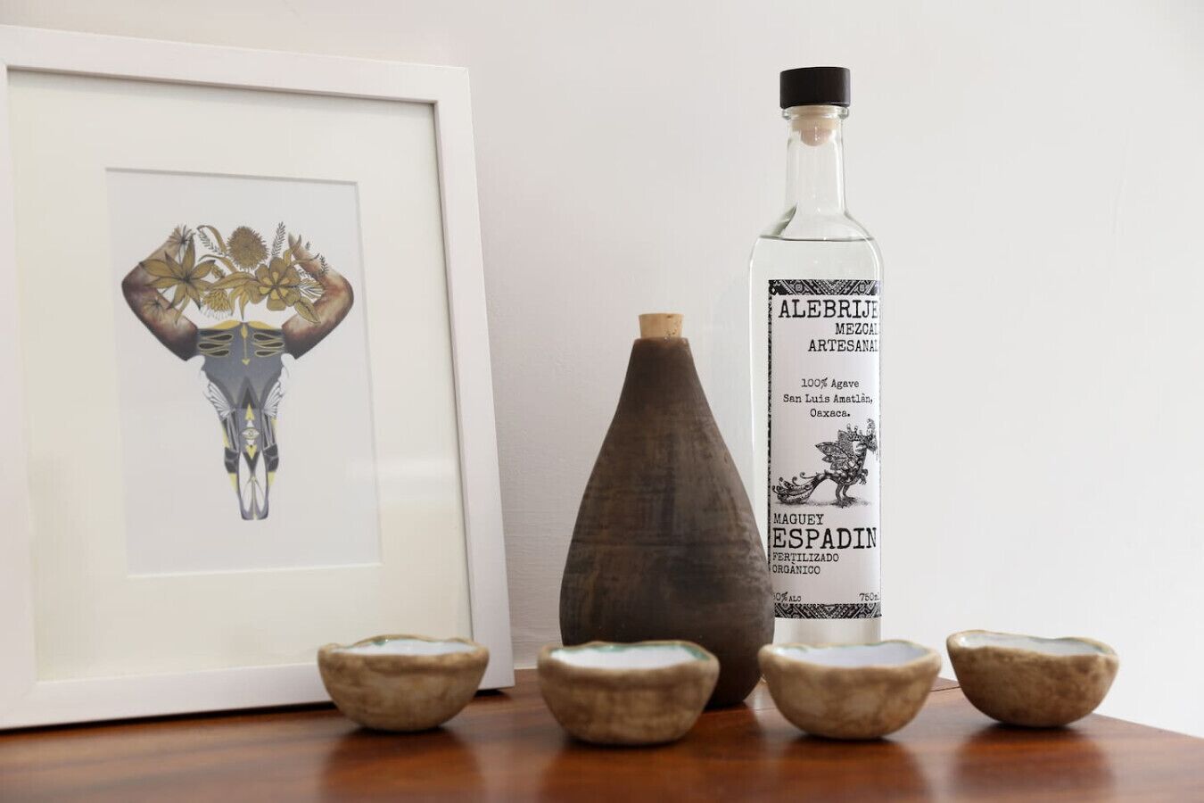 Une bouteille de mezcal Alebrije, une illustration botanique encadrée et quatre bols en céramique posés sur une surface en bois.