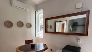 Une petite pièce bien éclairée, avec une table ronde en bois, une chaise, un petit tabouret, un miroir et un réfrigérateur.