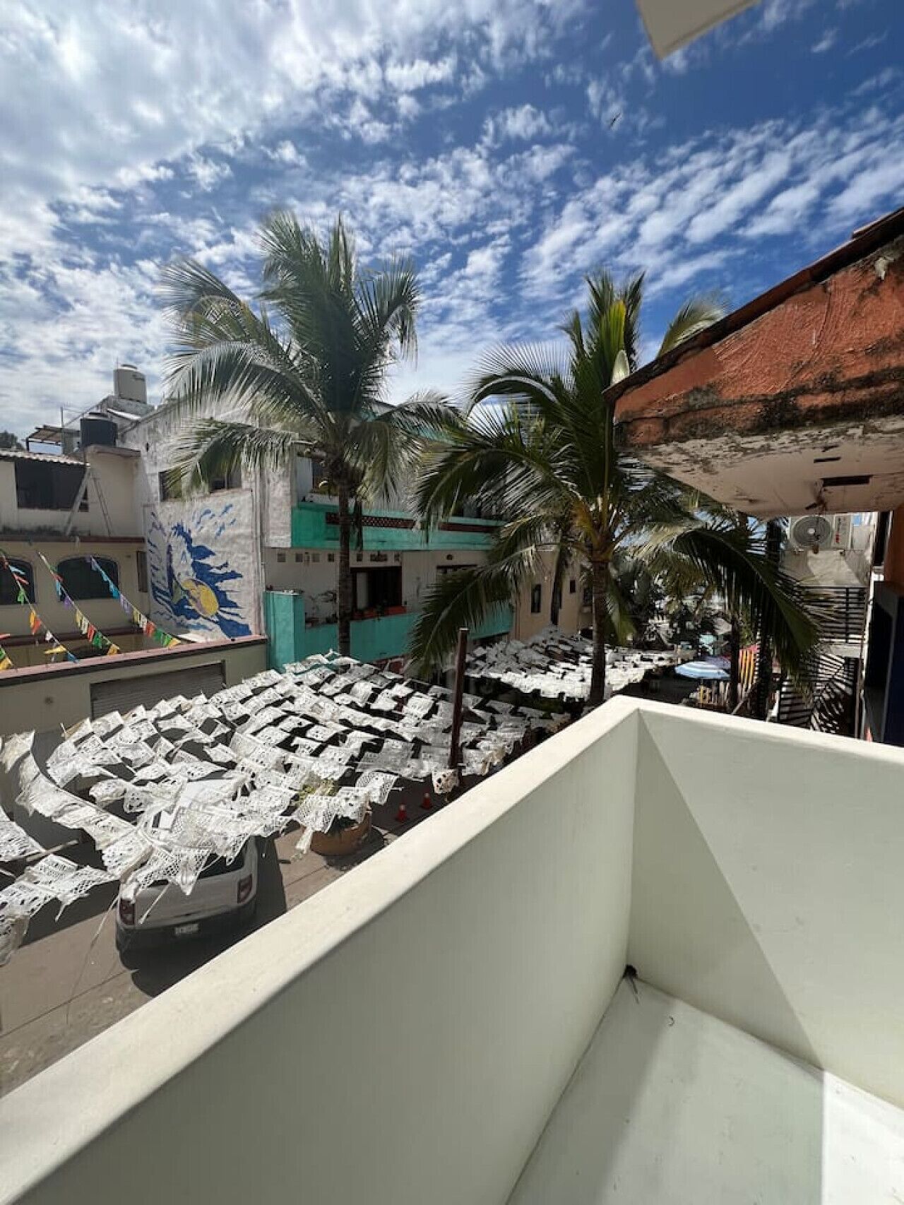 A balcony with a view of a courtyard covered with white cloths, palm trees, and buildings with colorful facades.