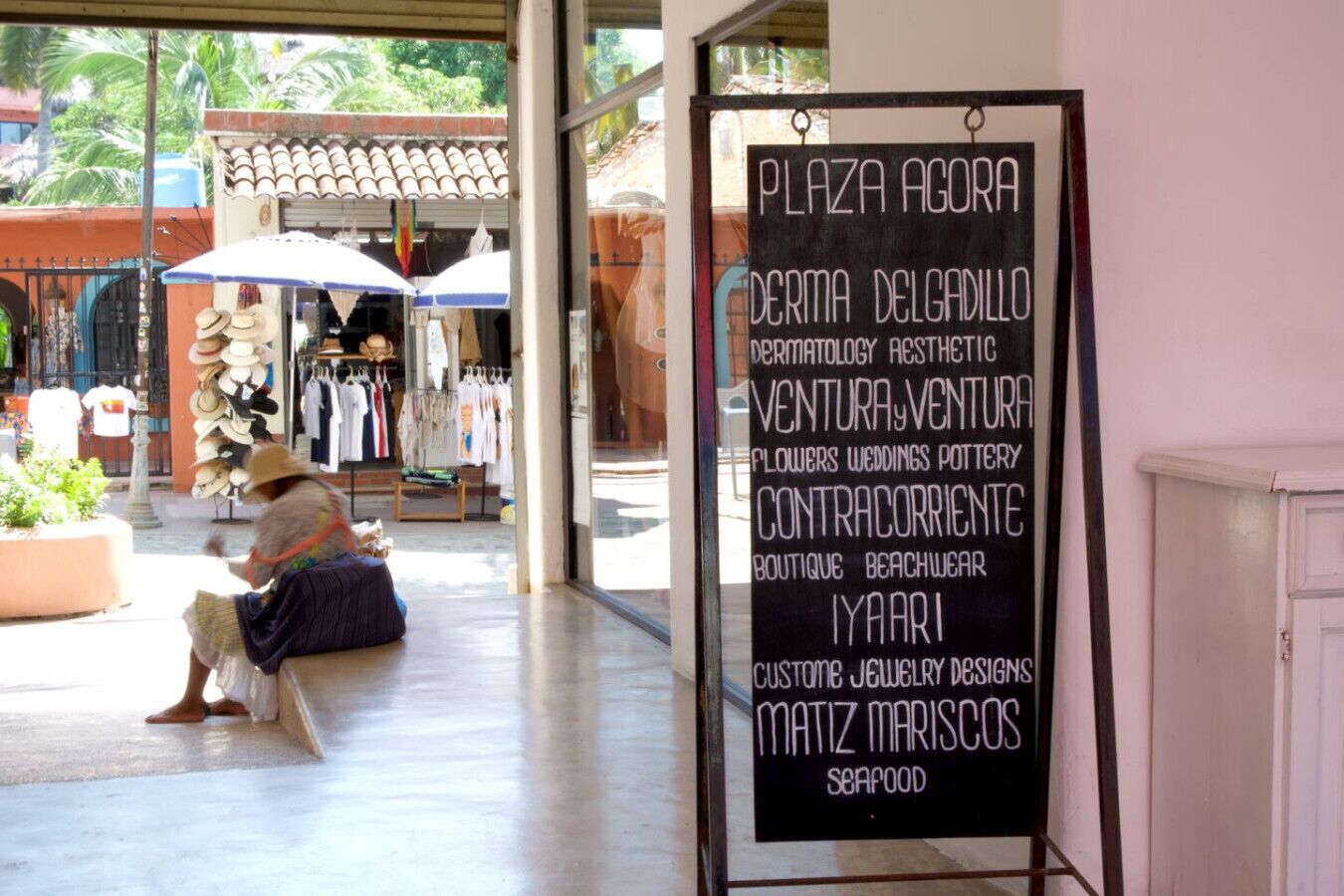 Un espace extérieur aménagé avec des sièges, où une personne est assise sur un banc, un panneau indiquant les noms de différents commerces, et une devanture de magasin où sont exposés des articles de vêtements.