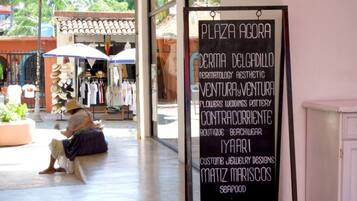 Un espace extérieur aménagé avec des sièges, où une personne est assise sur un banc, un panneau indiquant les noms de différents commerces, et une devanture de magasin où sont exposés des articles de vêtements.