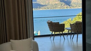 Quarto moderno com uma grande janela que oferece vista para um lago e montanhas. Há um sofá e duas cadeiras de vime com uma mesa.