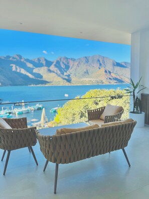 A balcony with two wicker chairs, a potted plant, and a view of a lake with boats and mountains. - Luxury Atitlan Suites (San Pedro La Laguna)