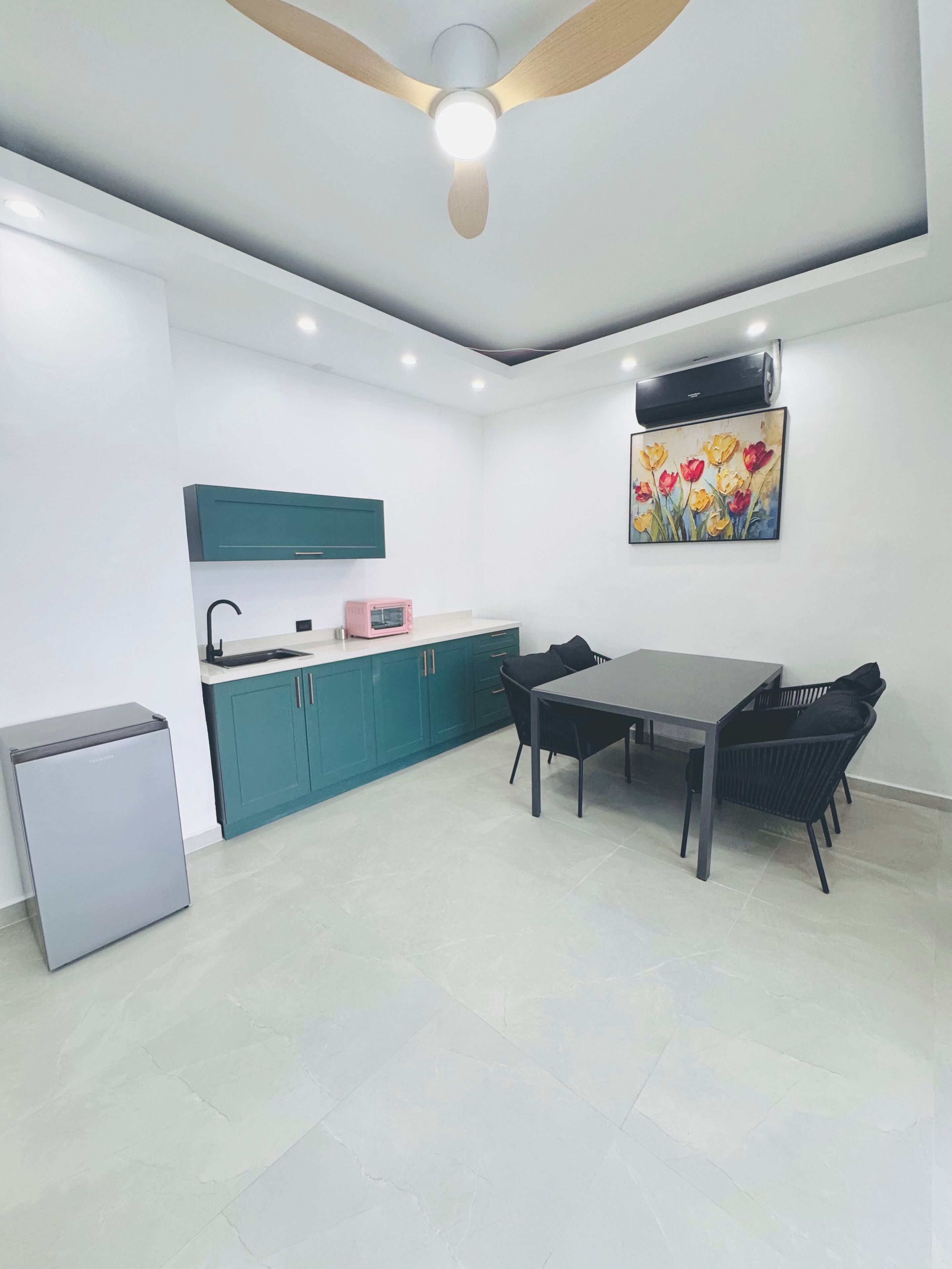 Una cocina compacta con gabinetes verde mar, una encimera blanca, un microondas rosa y un ventilador de techo.