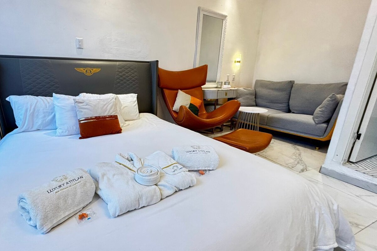 A hotel room with a large bed, a grey sofa, a brown armchair, and a small table with a lamp.
