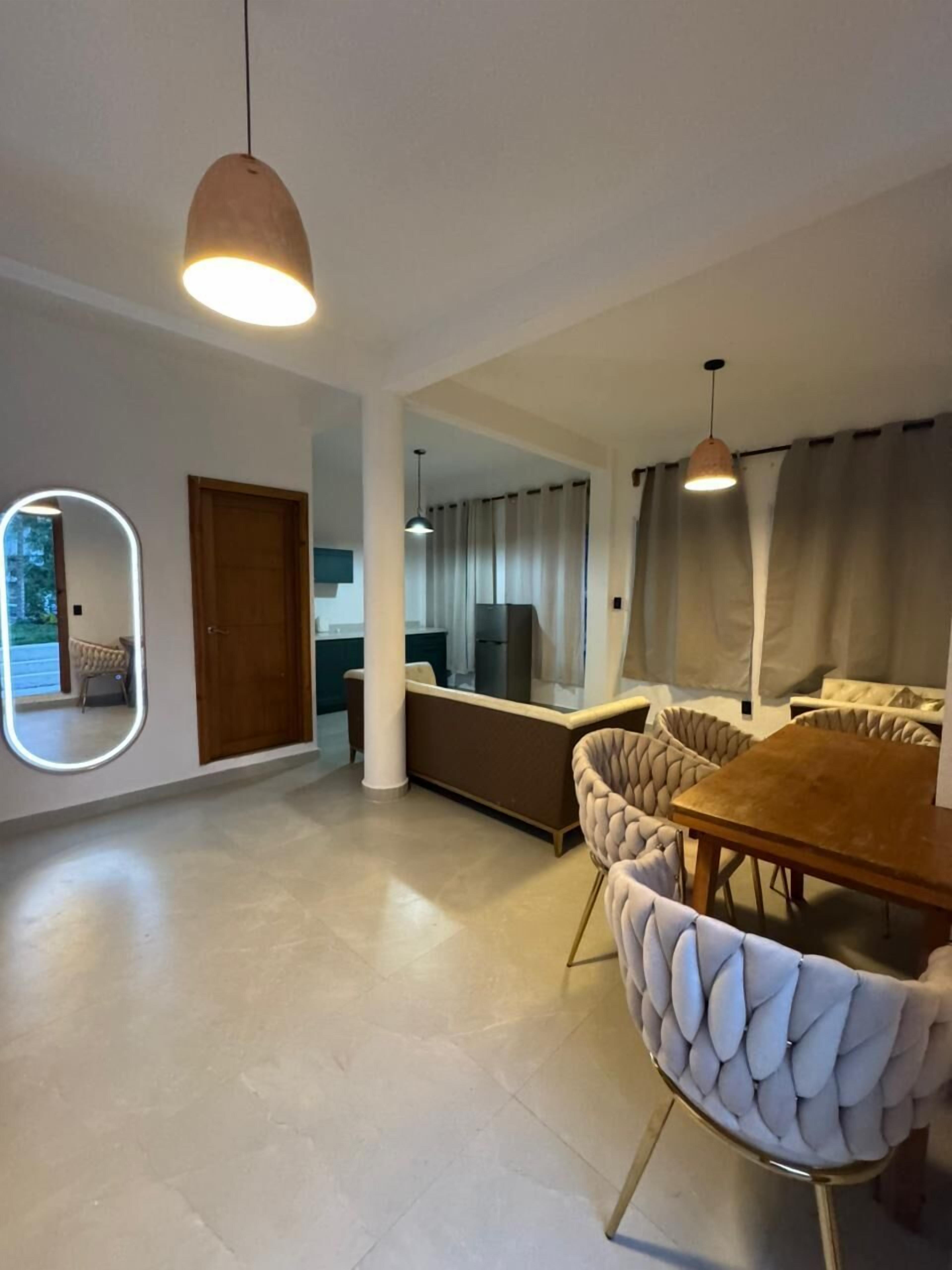 A modern living room with a dining area, featuring a wooden dining table, cushioned chairs, a sofa, and a large mirror.