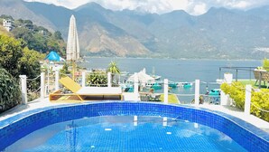 Piscina com azulejos azuis, rodeada por um deck com espreguiçadeiras e vista para montanhas e um lago.