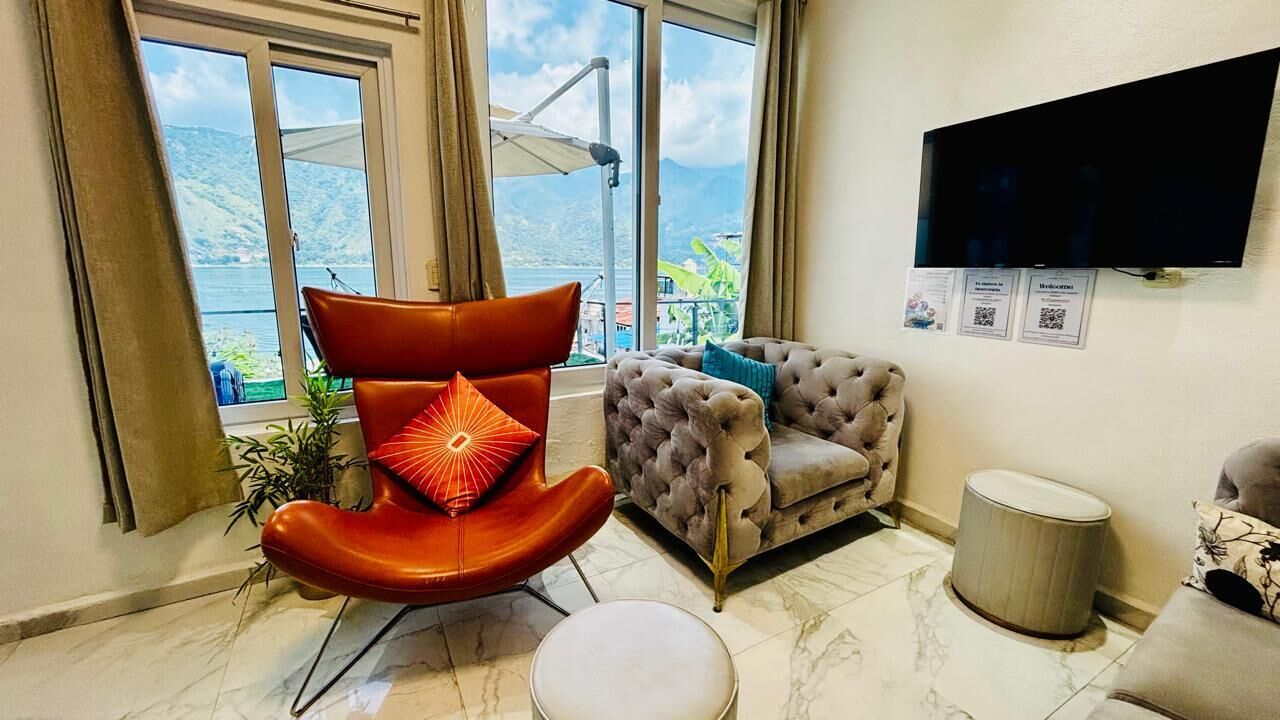 Una sala de estar moderna con un ventanal que ofrece vistas a un lago y montañas, un televisor de pantalla plana en la pared y una zona de estar cómoda con un sofá capitonado y una silla anaranjada original.