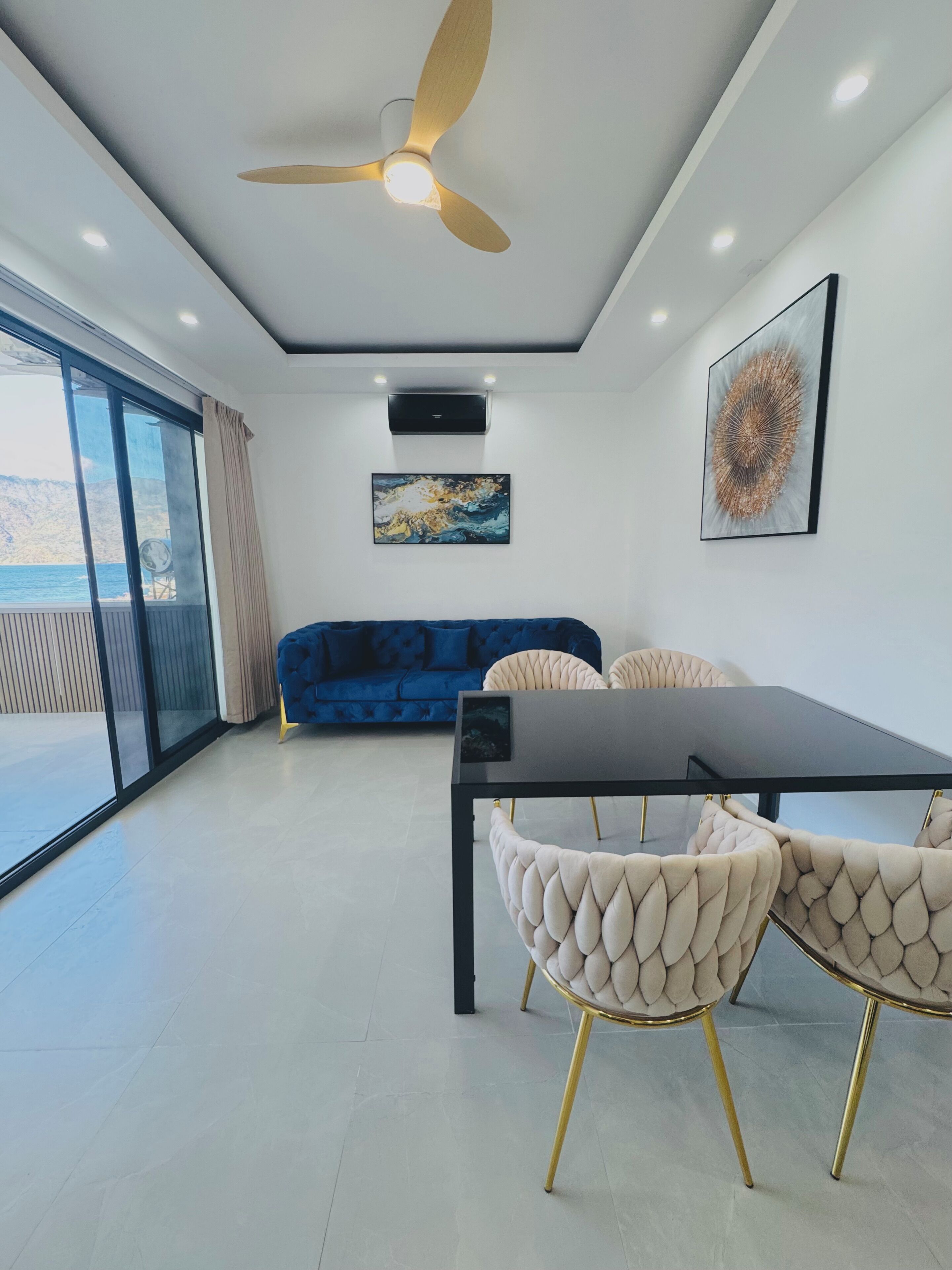 Una sala de estar moderna con un sofá azul, una mesa de comedor con sillas, un ventilador de techo y un ventanal con vistas al mar.