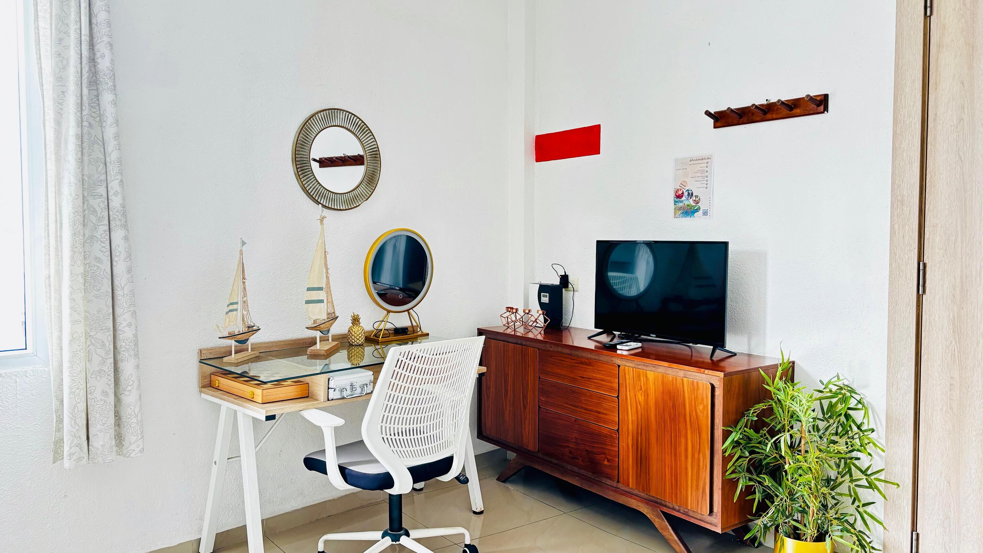 Un espacio habitable pequeño con un escritorio de madera, una silla de oficina blanca, un televisor, un espejo y una pequeña planta en maceta.
